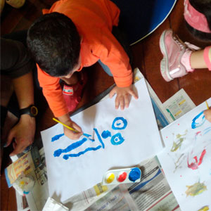 Children doing paintings, sitting on the floor, with papers and paints.