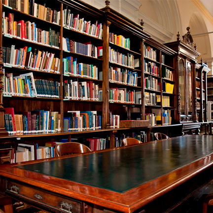 Uma estante repleta de livros e, à frente, uma grande mesa escura de madeira rodeada por cadeiras.