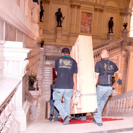 Homens transportando uma grande caixa de madeira. Eles descem as escadas do saguão. Ao fundo se vê uma parede com estátuas.