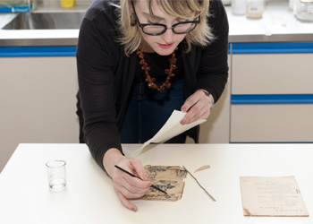 Uma mulher curvada sobre uma mesa. Ela utiliza um objeto semelhante a um pincel para restaurar uma obra de papel.
