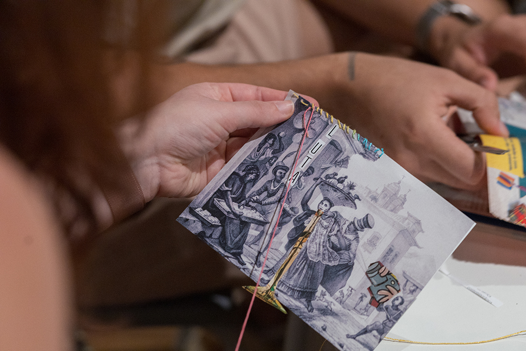 Mãos seguram uma imagem de Debret com colagens e costuras com linhas coloridas durante a oficina “O avesso de Debret: costurando e colando novas narrativas”, no Museu do Ipiranga