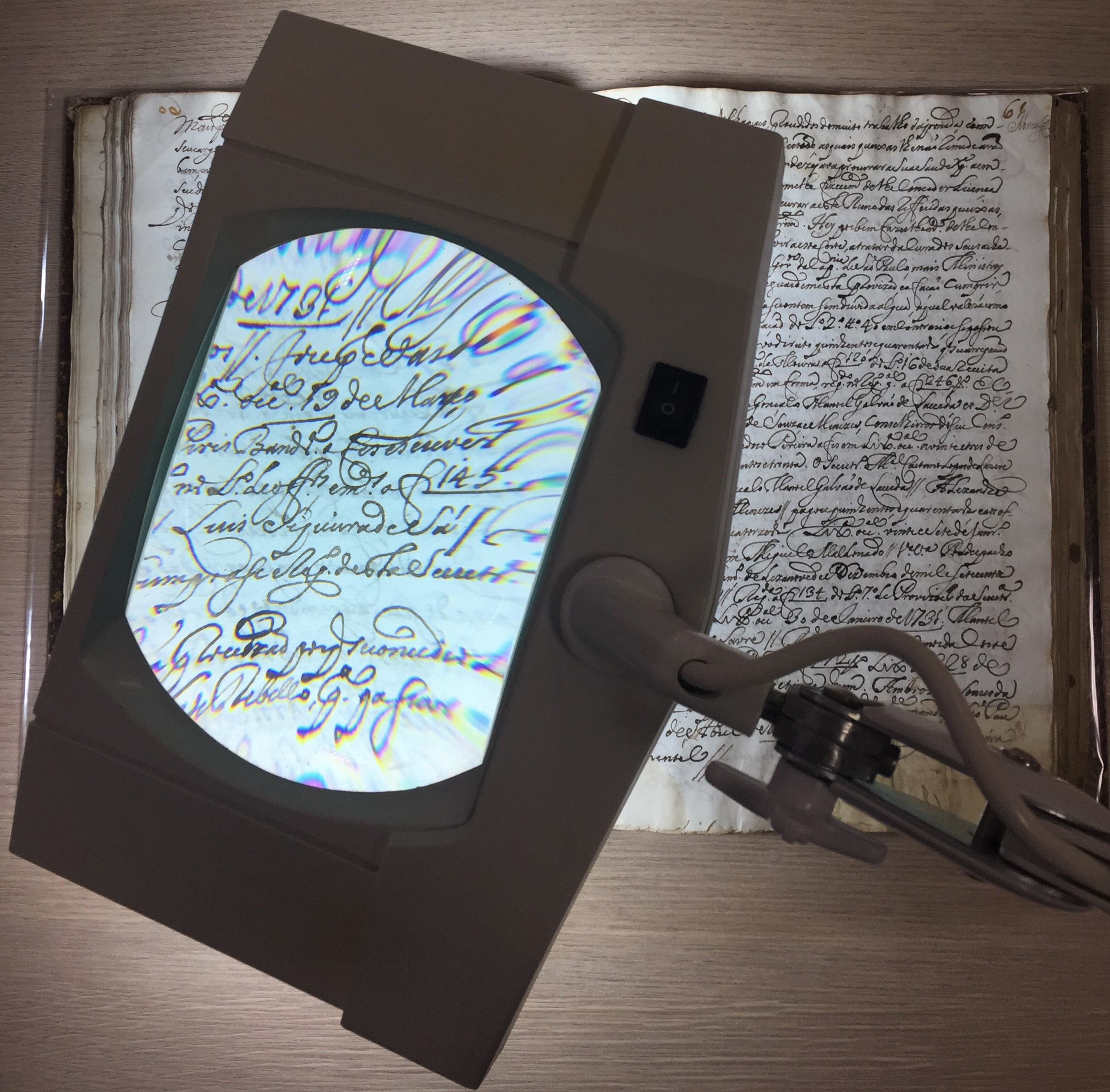 Lupa de aumento amplia a escrita manuscrita de um documento antigo aberto sobre a mesa, evidenciando detalhes do papel e da caligrafia