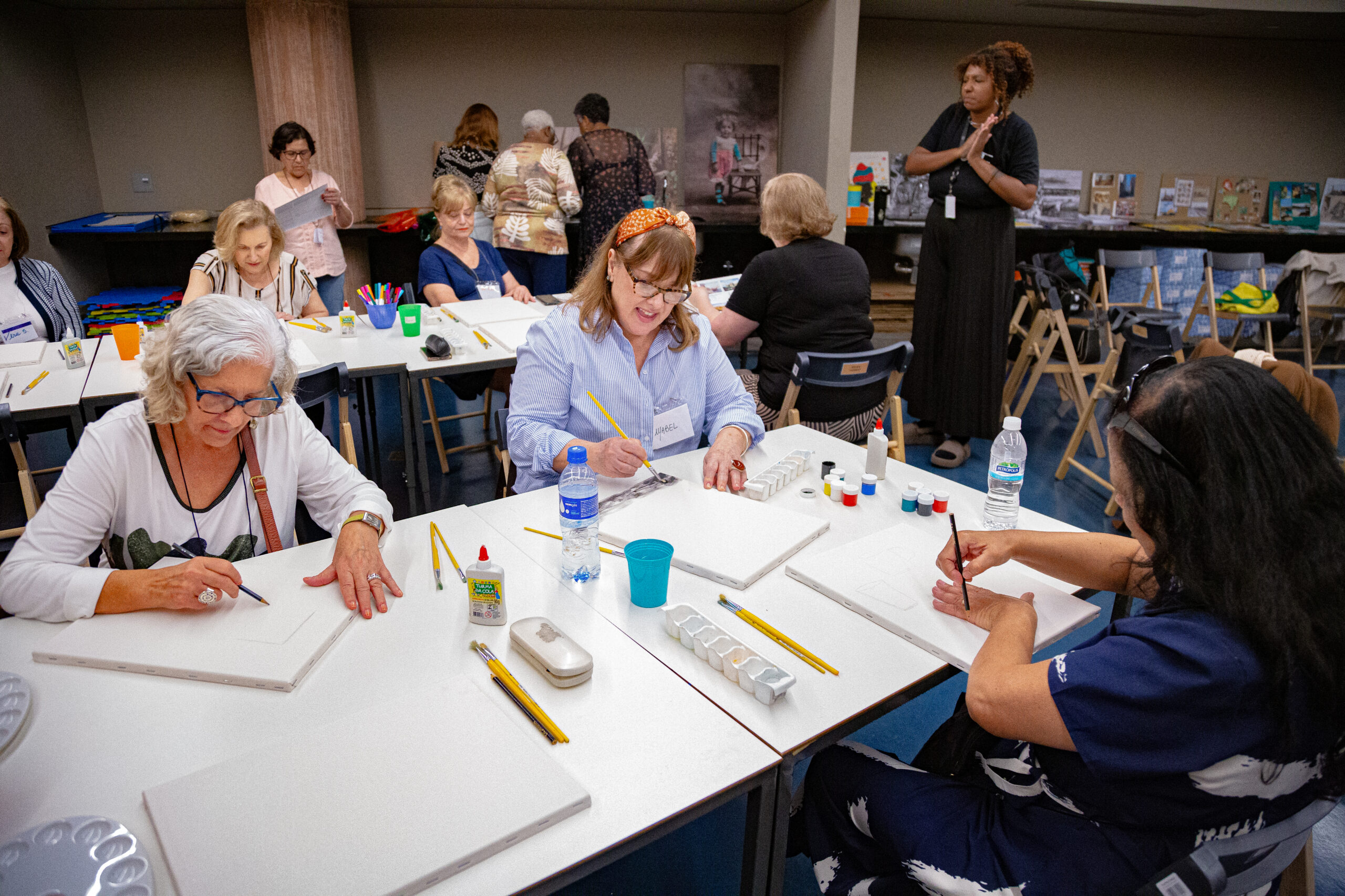 Participantes de uma oficina de pintura, em sua maioria mulheres idosas, estão sentadas ao redor de mesas, desenhando e pintando sobre telas brancas. Sobre as mesas, há pincéis, tintas, lápis e outros materiais artísticos. Ao fundo, uma educadora acompanha a atividade.